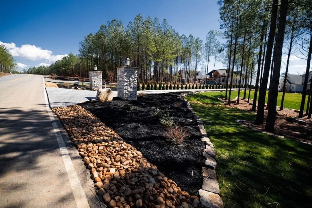 Stone entrance columns with river rock and mulch landscaping beside a road