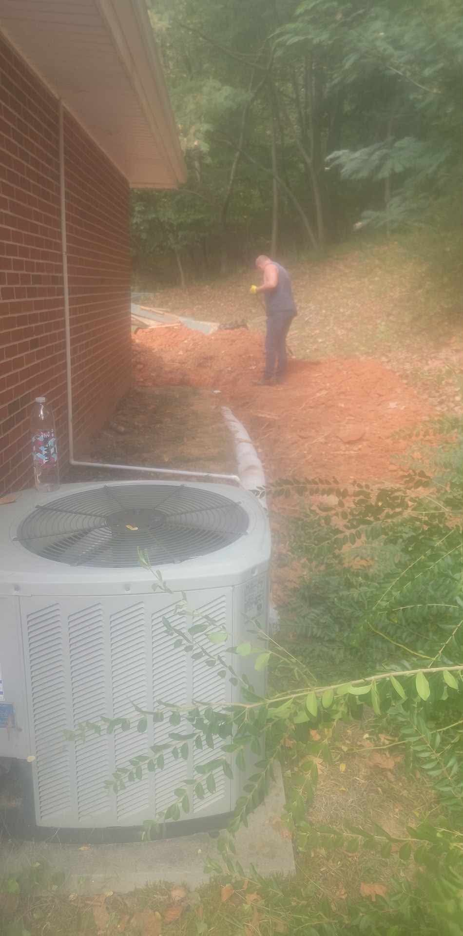 Worker grading red clay beside a brick house and air conditioning unit