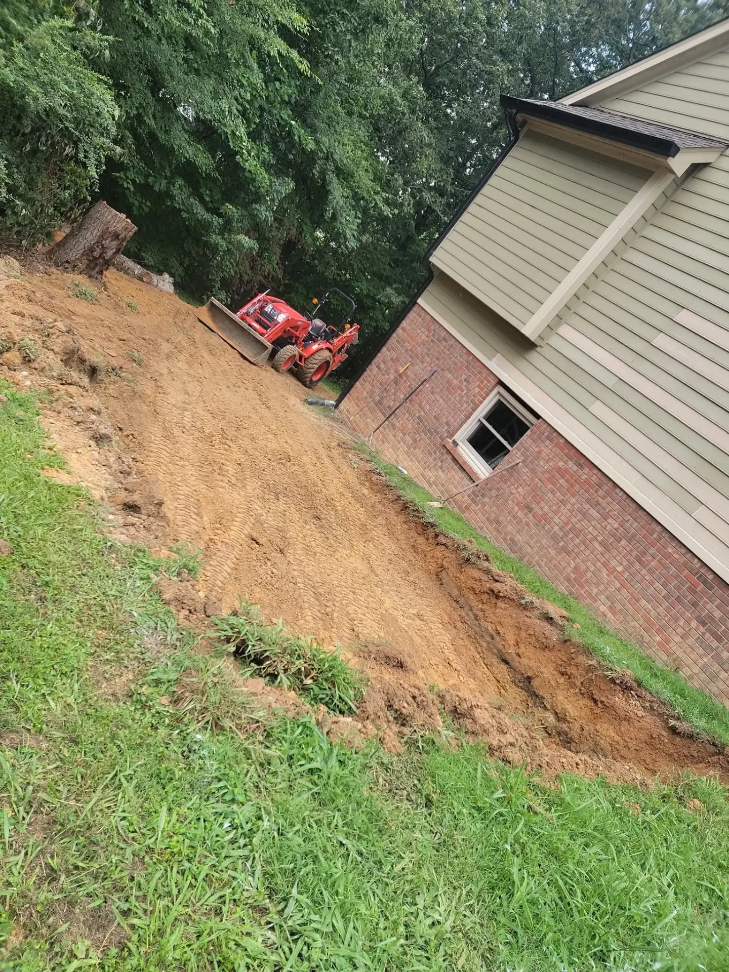 Red tractor grading a dirt slope beside a brick house