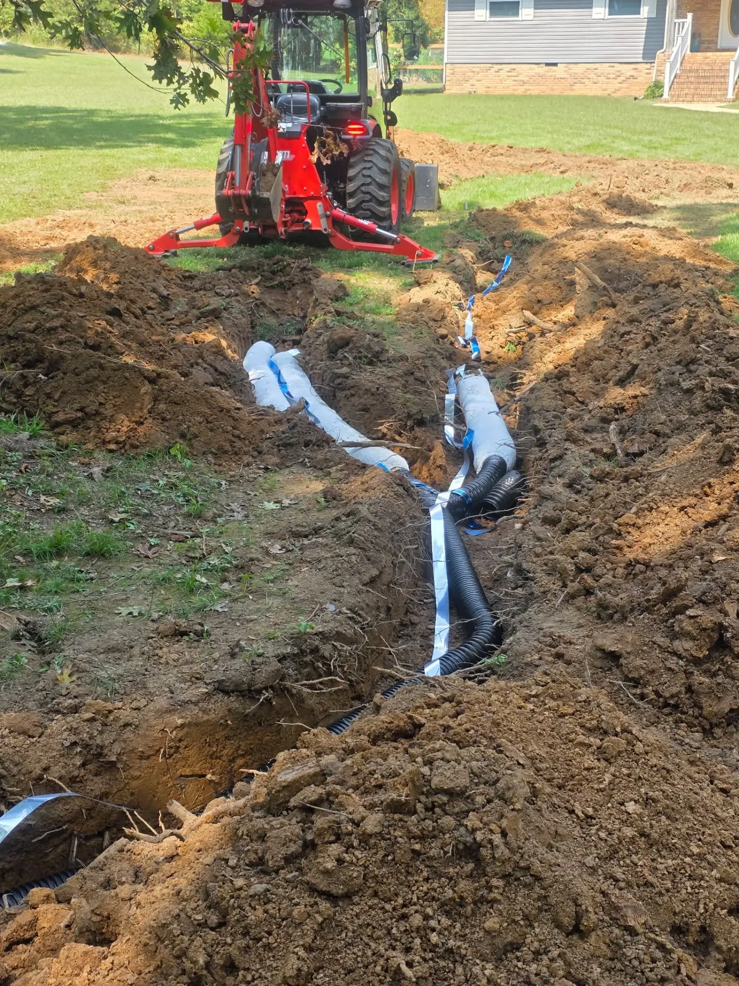 Backhoe behind a drainage trench with corrugated drain pipe and socked pipe laid in